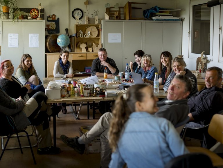 12 Menschen sitzen um einen großen Tisch in einer Werkstatt, unterhalten sich und lachen.