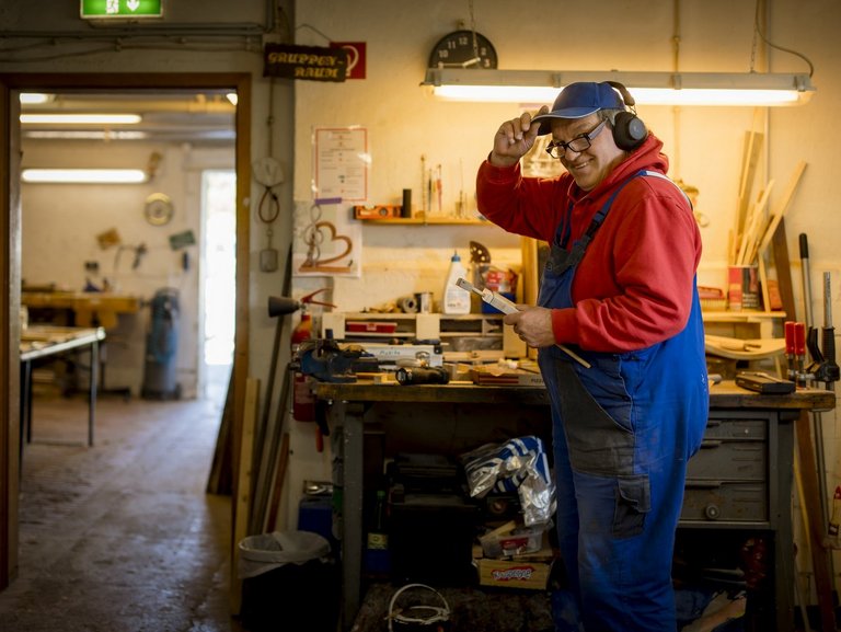 Ein Mann im blauen Overall und roten Pullover steht vor einem Arbeitstisch in der Werkstatt und tippt sich zum Gruß an die Mütze.