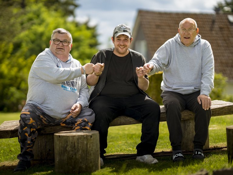 Drei Männer sitzen auf einer Holzbank und machen lachend einen Faustgruß.